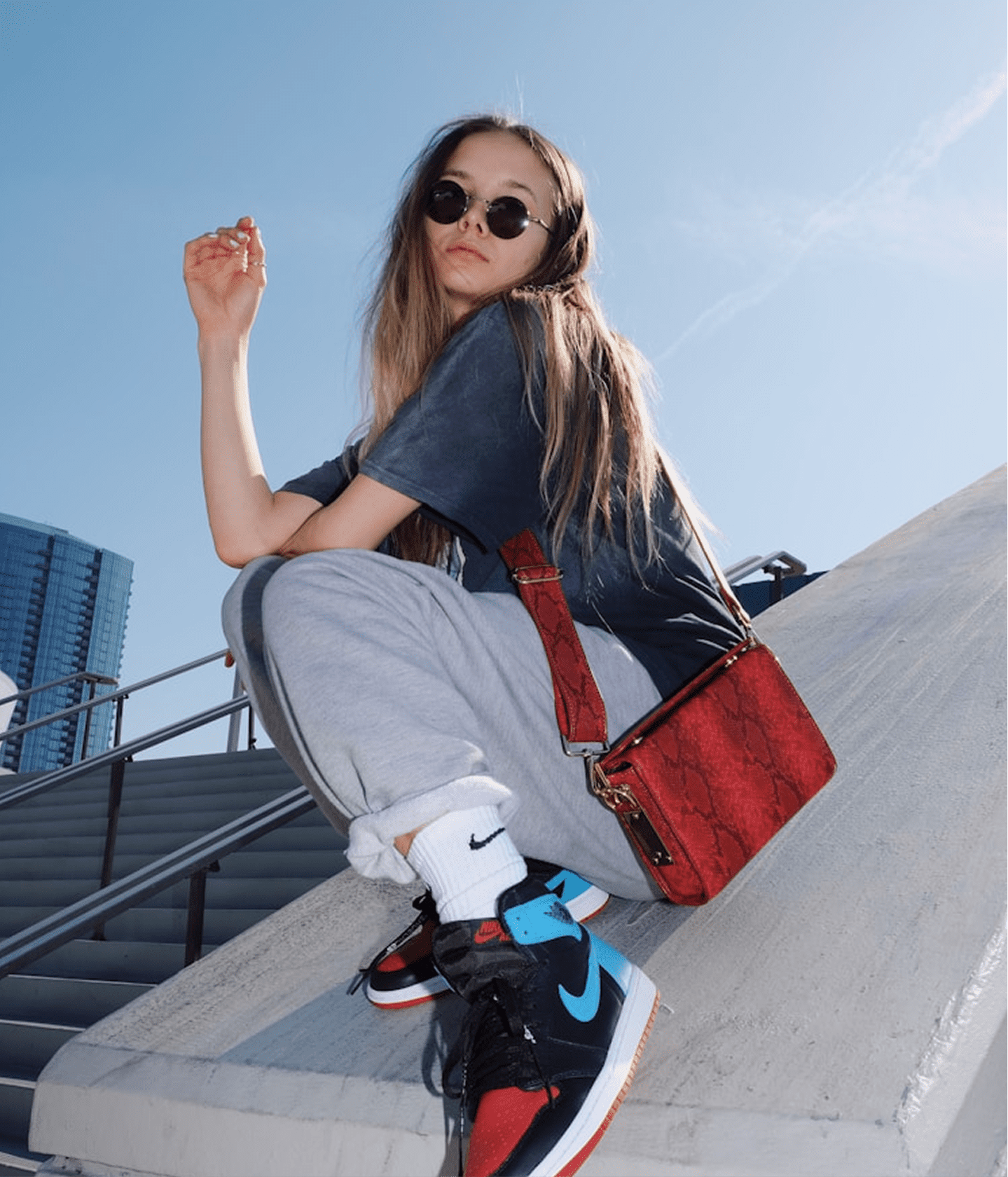 A woman wearing round sunglasses, a dark gray t-shirt, light gray sweatpants, and a red textured shoulder bag sits on concrete stairs outdoors under a blue sky. She is wearing black and red high-top sneakers with a bright blue Nike Swoosh.