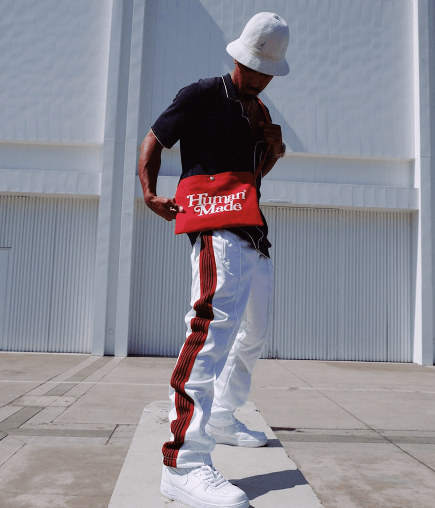 A person wearing a white bucket hat, black polo shirt, white pants with red side stripes, and white sneakers stands outdoors in front of a white building, holding a red bag with 'Hungry Made' printed on the front.