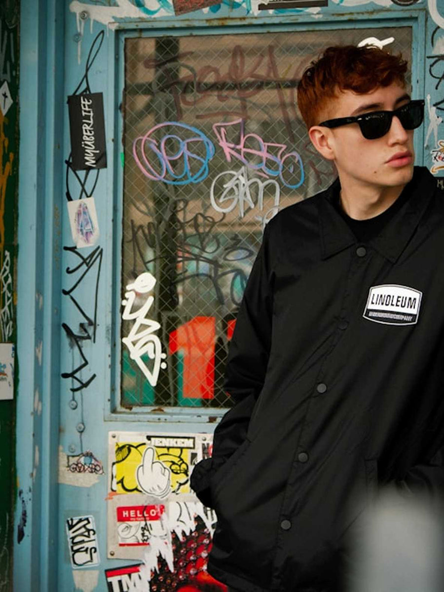 A young man with reddish-brown hair and sunglasses wearing a black Linoleum jacket, standing in front of a graffiti-covered blue door.