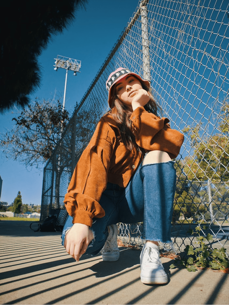A person crouching outdoors in front of a chain-link fence on a sunny day, wearing a rust-orange cropped hoodie, blue jeans, white sneakers, and a pink bucket hat.