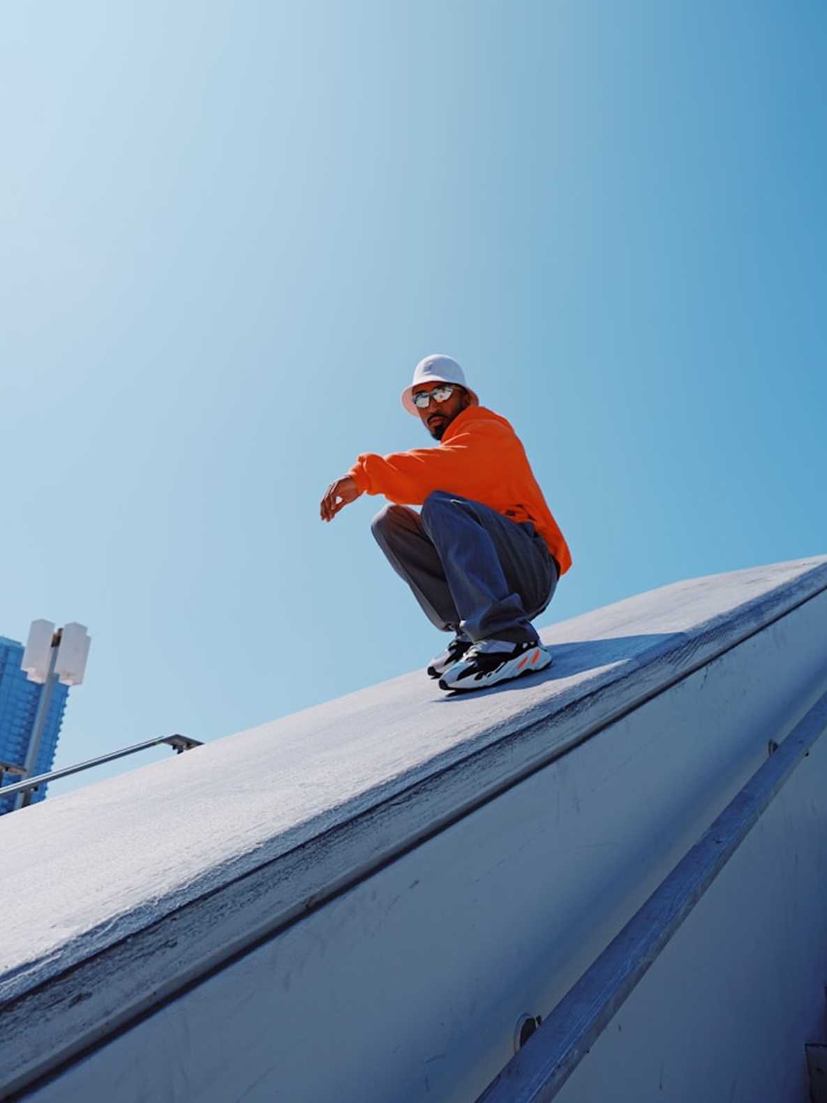 A person crouching on a skateboard on a white sloped roof under a clear blue sky, wearing an orange hoodie, gray pants, a white helmet, and black-and-white sneakers.