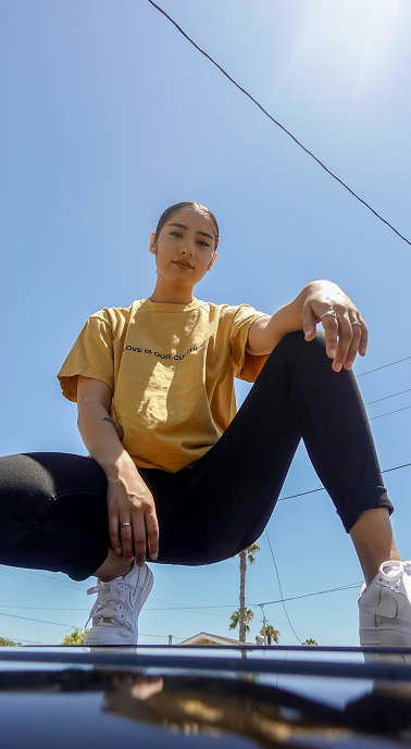 A woman wearing a mustard yellow graphic t-shirt, black pants, and white sneakers crouches on the hood of a car against a bright blue sky.