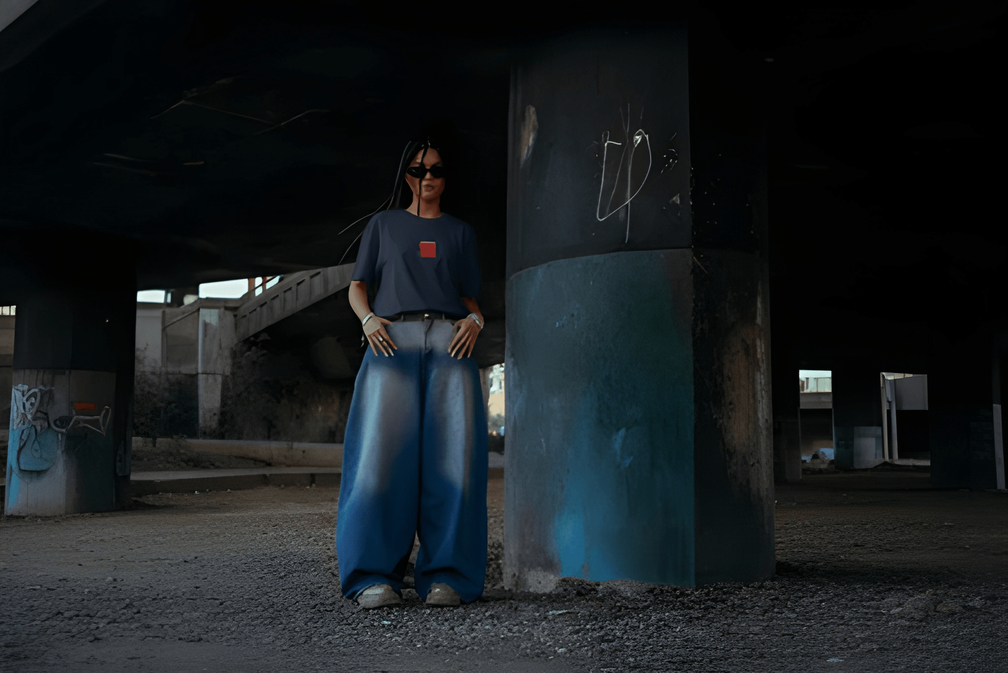 A person stands under a concrete bridge wearing a dark blue t-shirt with a small red square logo on the chest, wide-leg blue jeans, and sunglasses.
